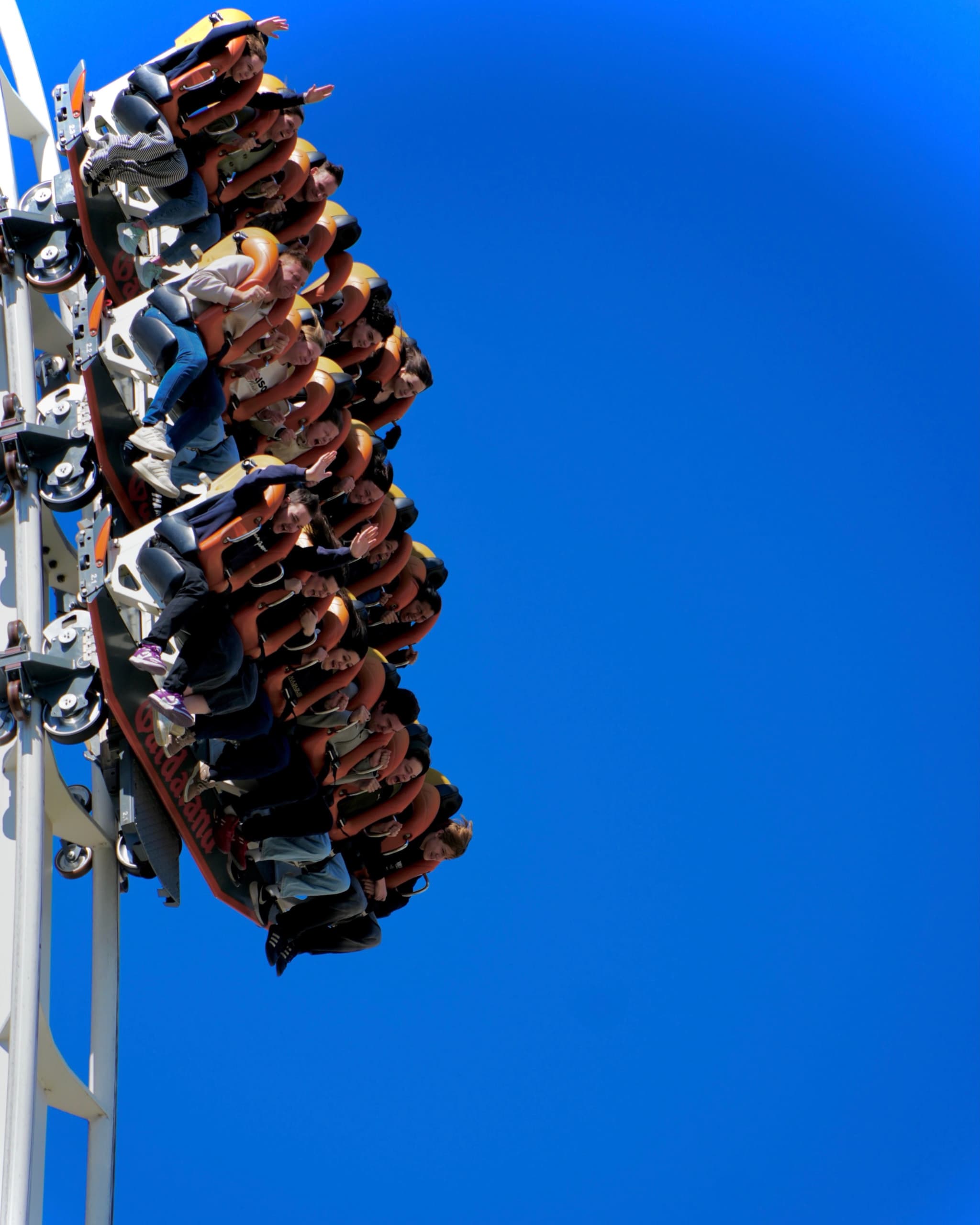 riders in orange seats ascend a vertical roller coaster track against a clear blue sky during a bright sunny afternoon at park