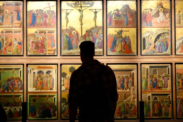 a visitor stands before illuminated religious panels in a dim gallery, observing gold-leaf paintings under soft overhead museum lighting during the day