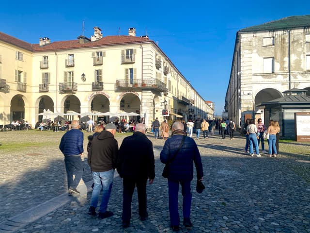 people walk across a cobblestone plaza in front of historic buildings under a clear blue sky on a bright sunny day