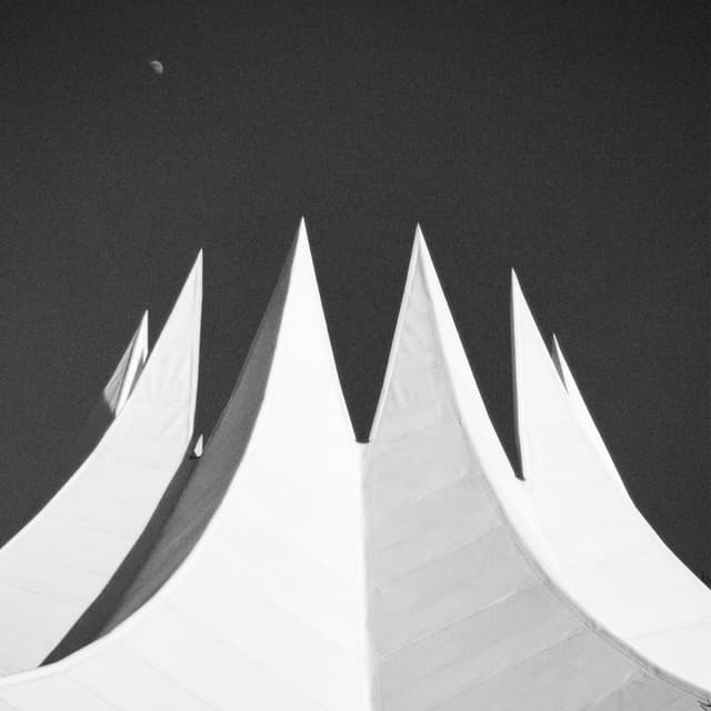Minimalist black and white architectural photography captures sharp, triangular peaks of a modern structure against a dark sky with a crescent moon