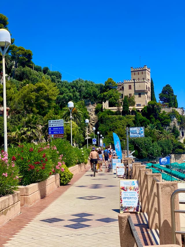 pedestrians walk along a paved coastal path toward a stone castle under a clear blue sky in bright midday summer sunlight