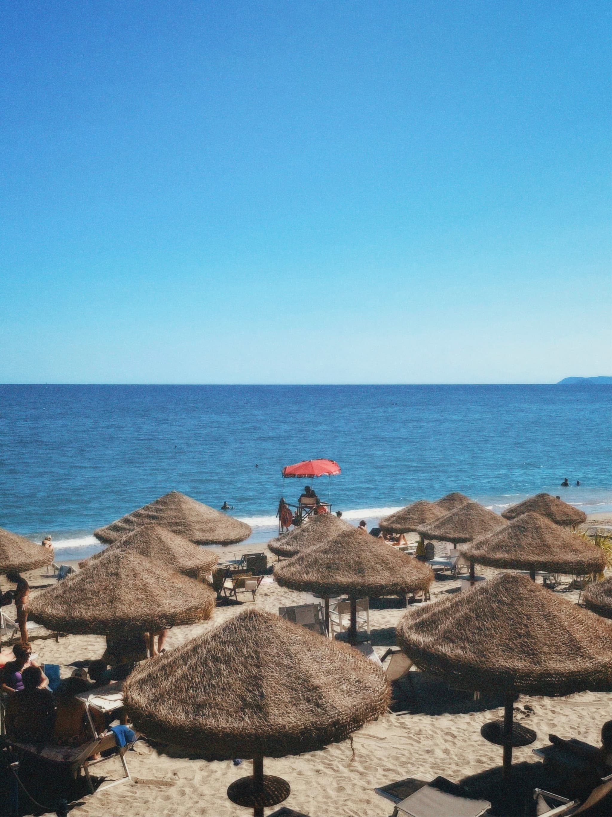 straw umbrellas shade a sunny beach with blue ocean water under a clear sky, featuring one red umbrella in the distance