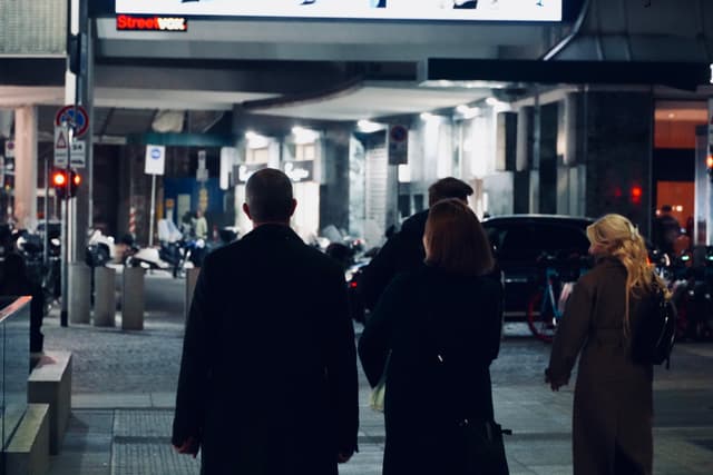 three people walk down a city street at night under bright overhead lights with cars parked along the concrete sidewalk
