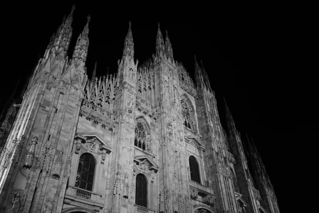 milan cathedral facade lit by night floodlights, showing gothic stone spires and dark arched windows in a black and white photograph