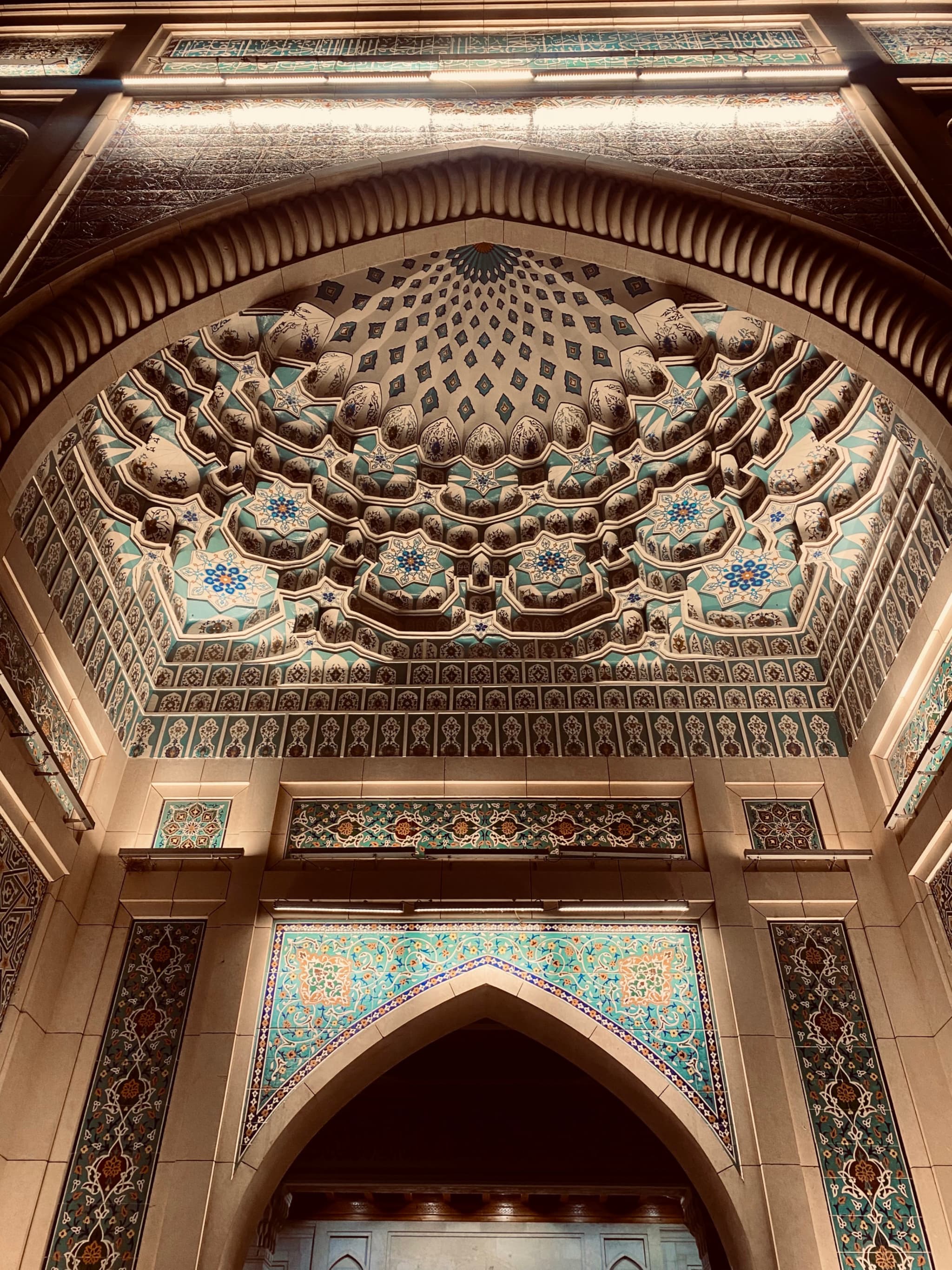 intricate muqarnas ceiling detail above a stone archway in a dimly lit mosque, featuring blue tile patterns and warm architectural lighting
