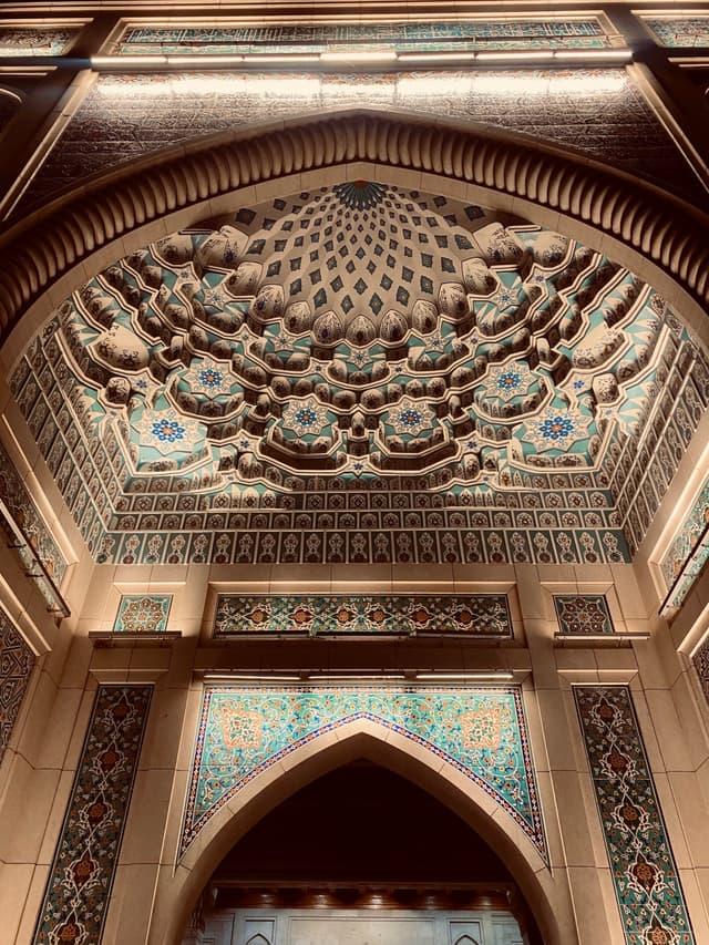 intricate muqarnas ceiling detail above a stone archway in a dimly lit mosque, featuring blue tile patterns and warm architectural lighting