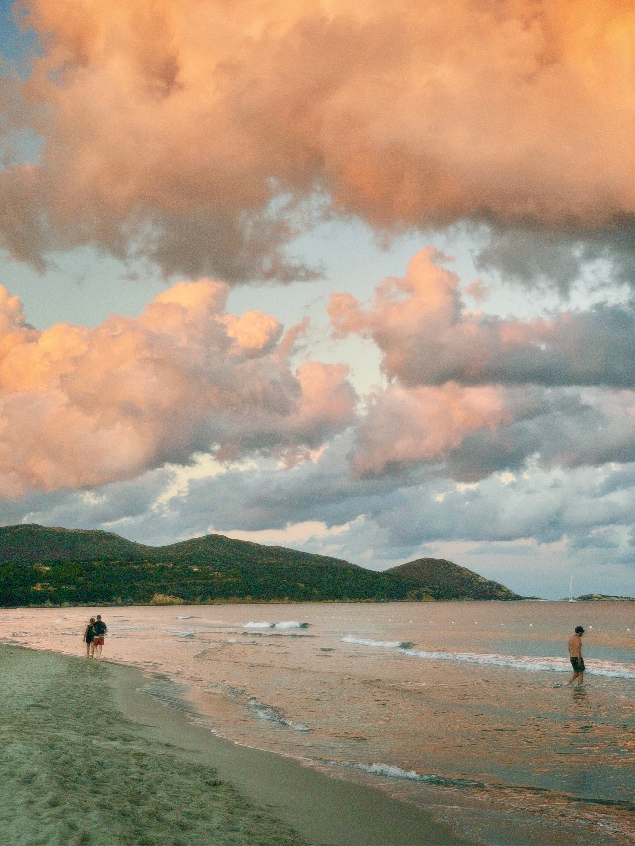 people walk along a sandy beach under orange sunset clouds with distant green hills and calm water during the late evening