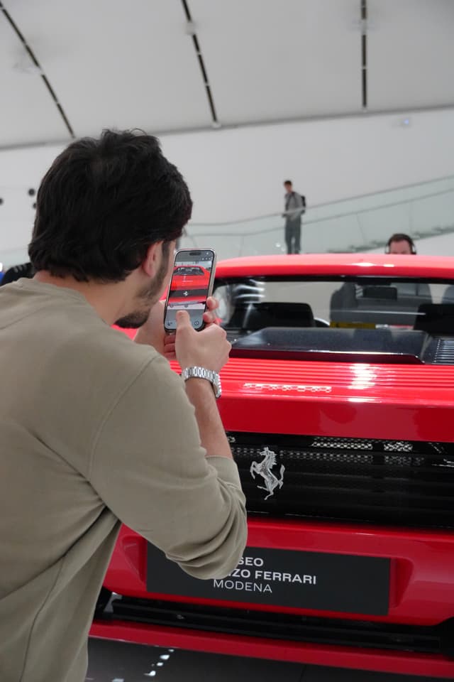man in beige sweater uses smartphone to photograph red ferrari sports car inside bright museum gallery with white walls and overhead lights