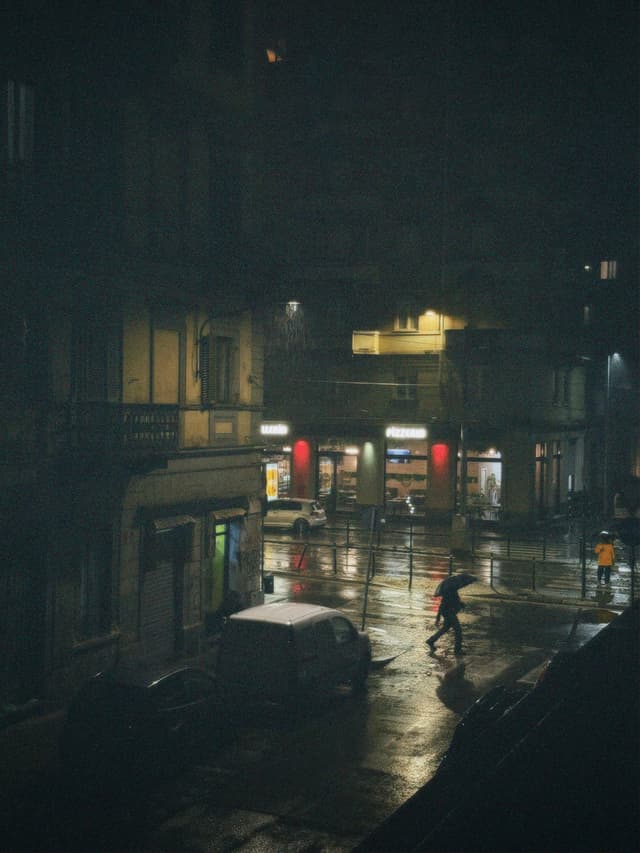 a person with an umbrella walks across a dark city street during rain under dim yellow artificial lights at night