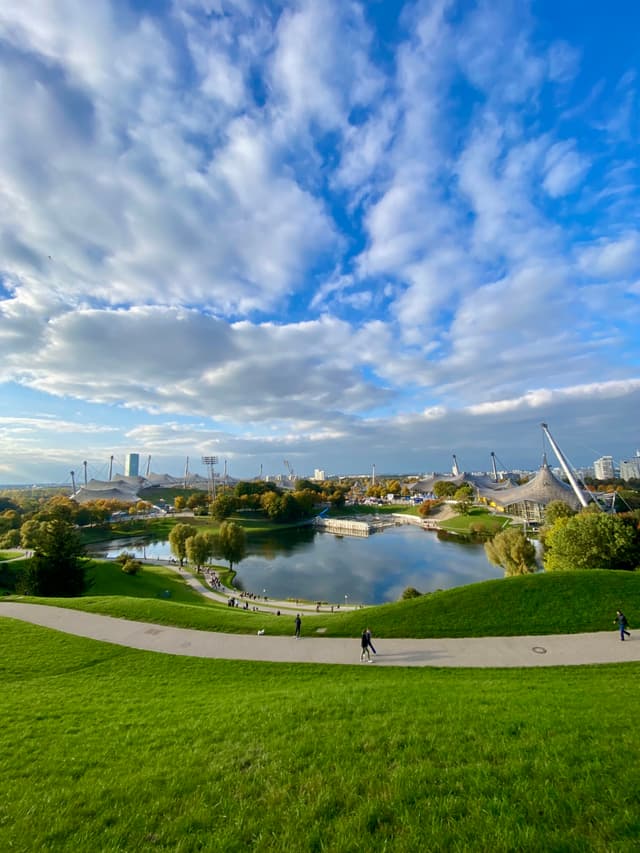 green hills overlook the olympic park lake in munich under a bright blue sky with scattered clouds on a clear afternoon