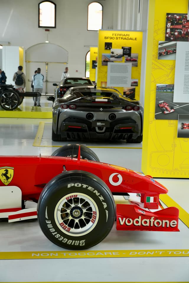 red ferrari formula one car parked in a bright museum gallery with a grey supercar and yellow information panels in the background
