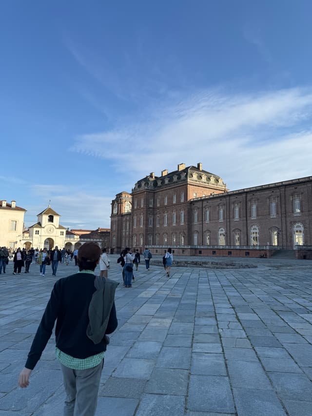 tourists walk across the stone courtyard of the royal palace of venaria in italy under a clear blue sky during the afternoon