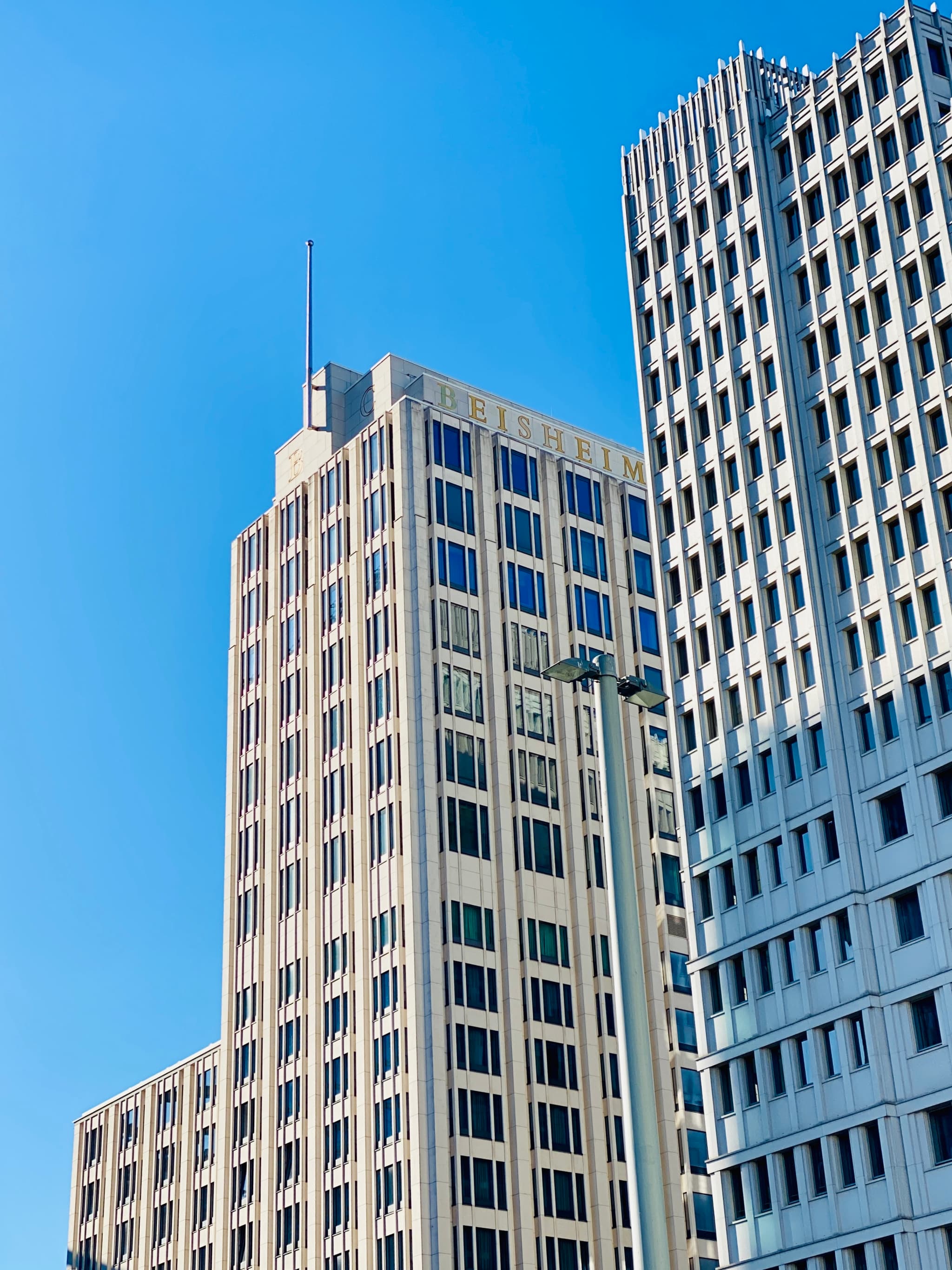 tall concrete office buildings rise against a clear blue sky under bright daylight, showing geometric window patterns and modern urban architecture