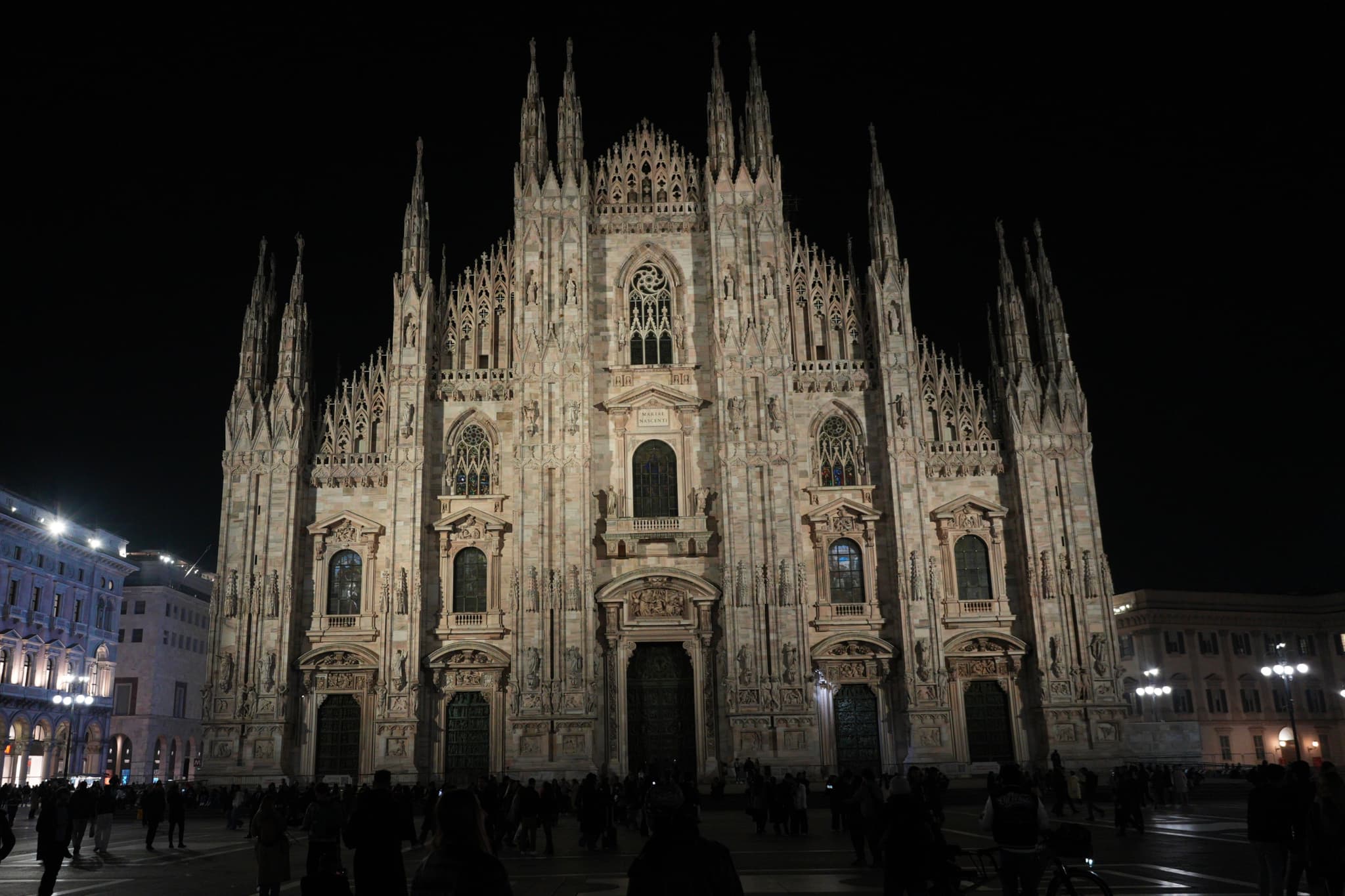 milan cathedral facade illuminated at night with sharp architectural details and dark silhouettes of people walking across the empty paved square