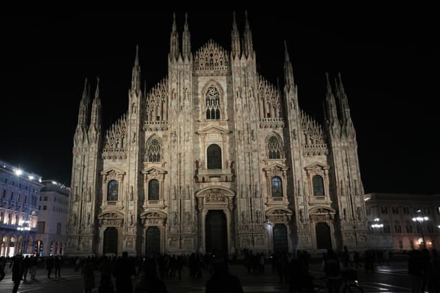 milan cathedral facade illuminated at night with sharp architectural details and dark silhouettes of people walking across the empty paved square