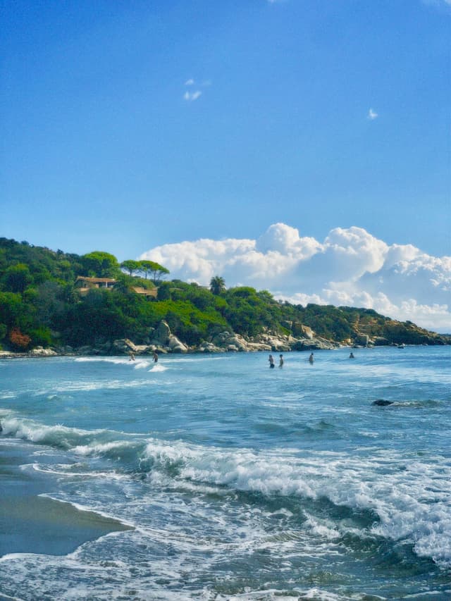 people swim in blue coastal waters under a clear sky with white clouds near a green hillside on a sunny day
