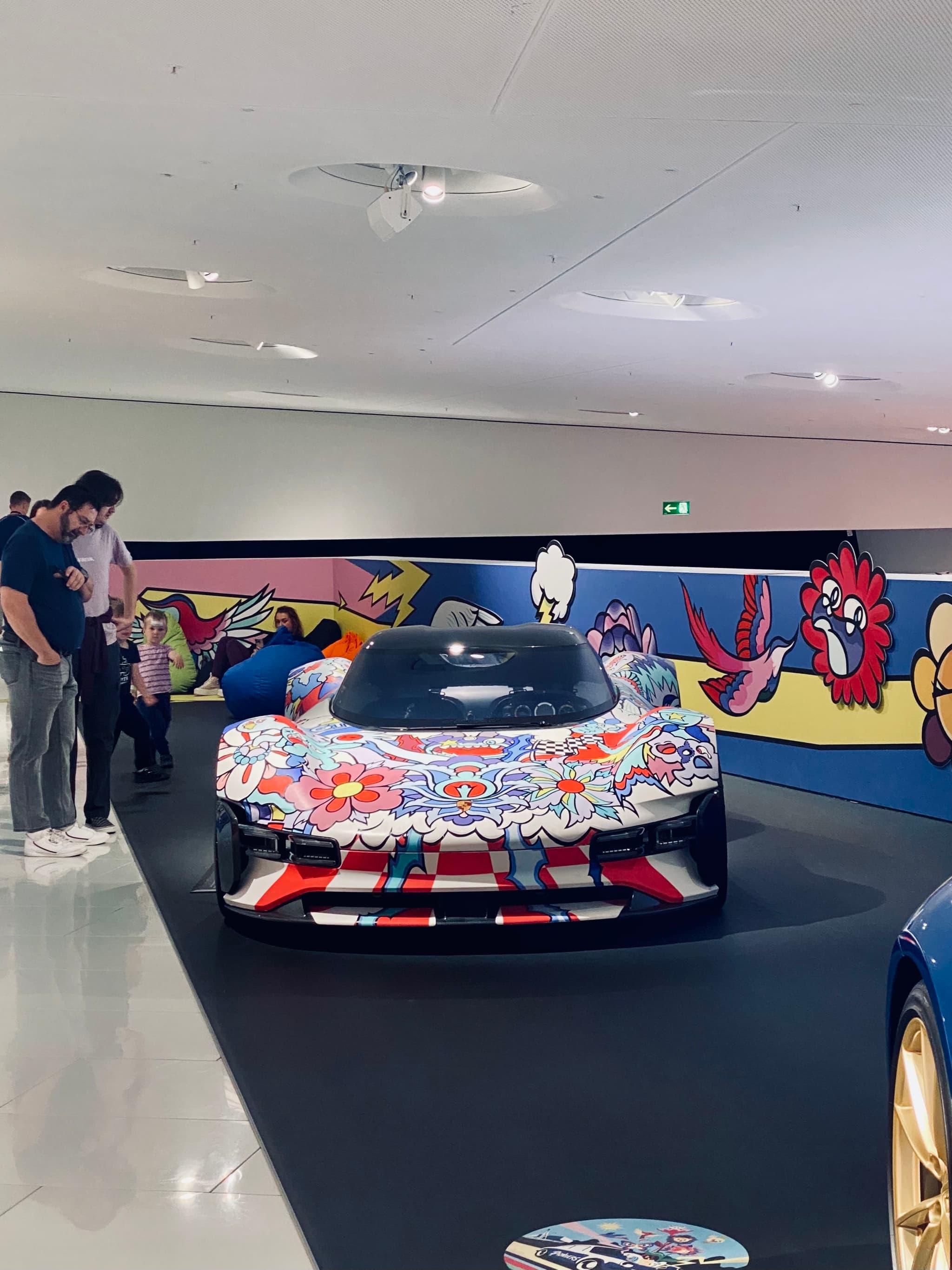 visitors view a colorful art car inside a bright museum gallery with painted wall murals and overhead spotlights on the floor