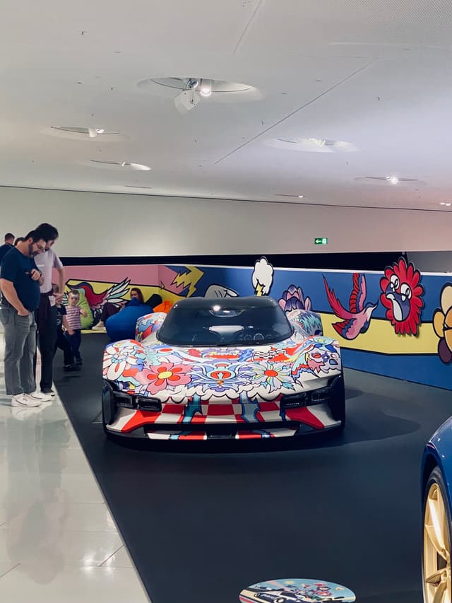 visitors view a colorful art car inside a bright museum gallery with painted wall murals and overhead spotlights on the floor