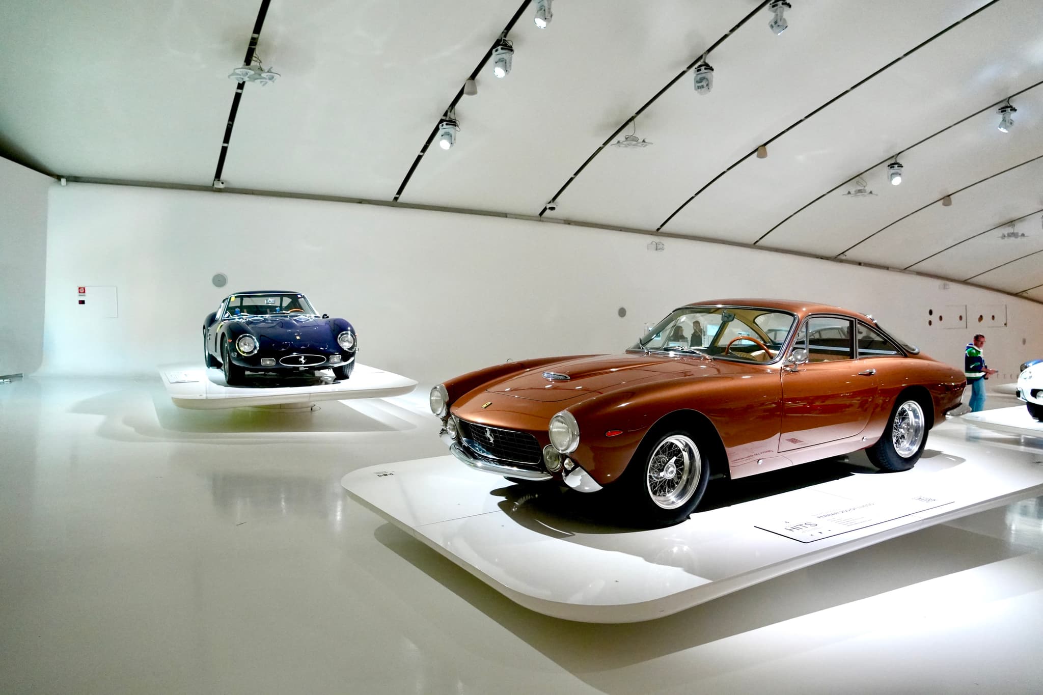 vintage copper and dark blue sports cars displayed on white platforms inside a modern museum with bright overhead track lighting