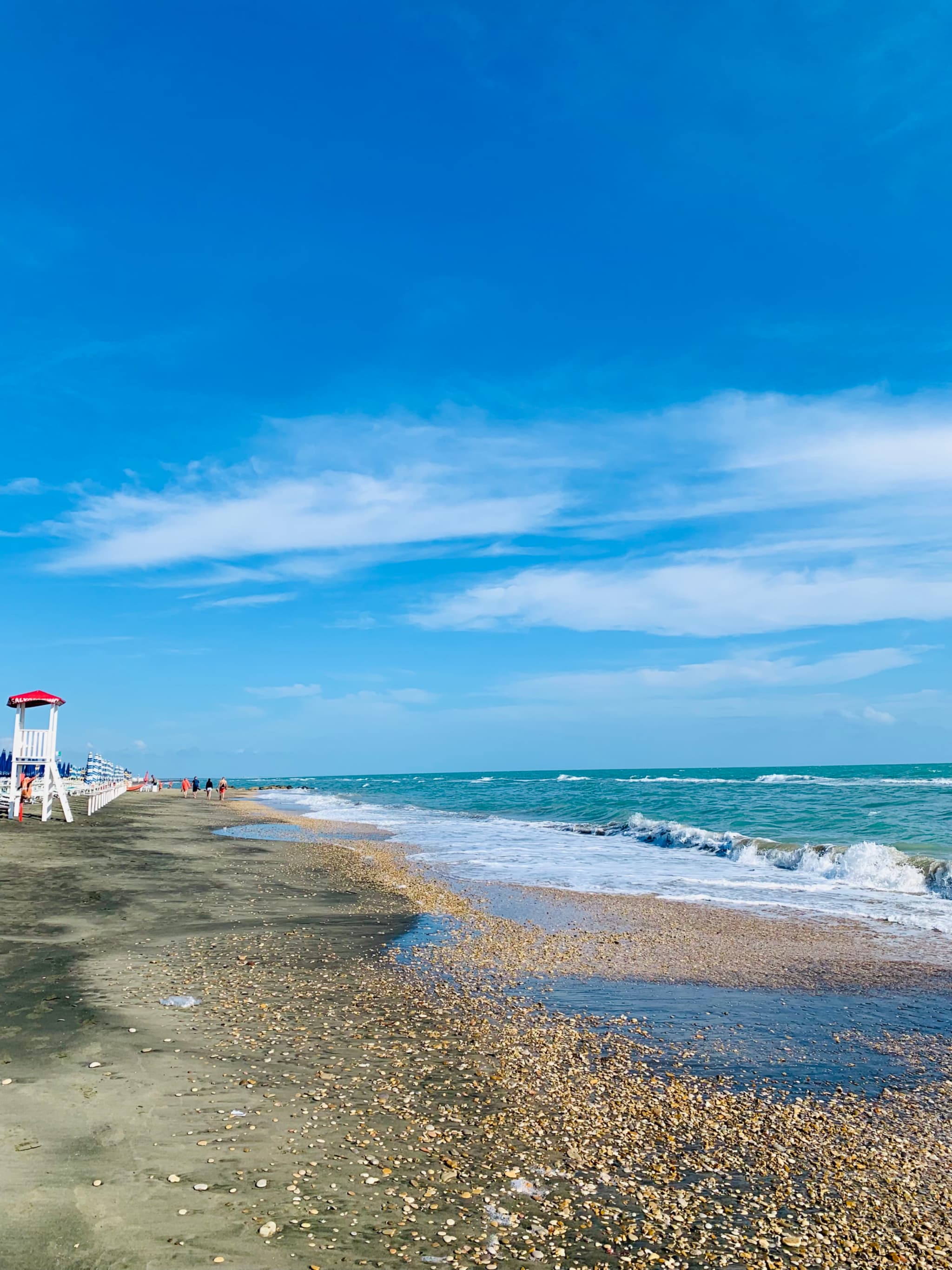 white lifeguard chair stands on a sandy beach under a bright blue sky with small waves hitting the shore in daylight