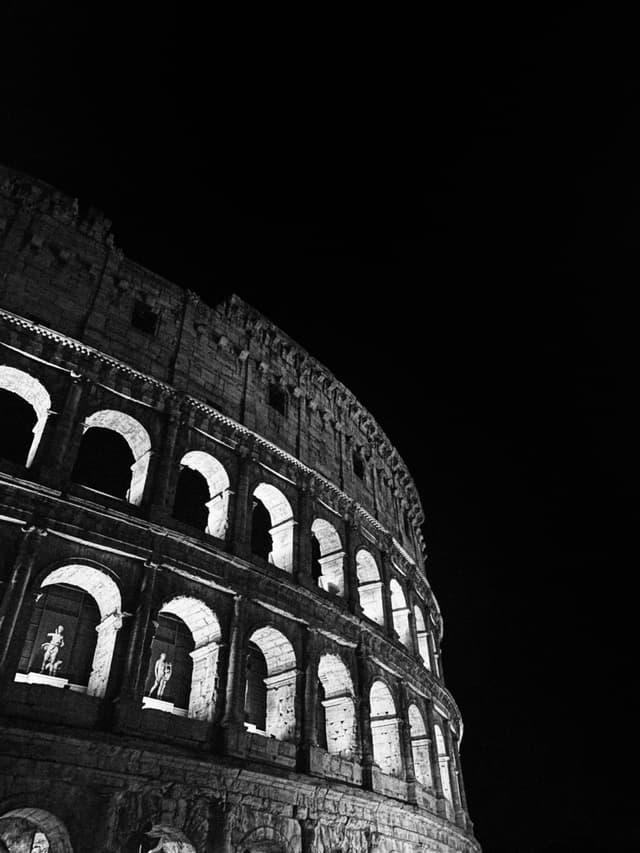 Dramatic black and white photography of the ancient Roman Colosseum illuminated at night, highlighting iconic architectural arches against a dark sky