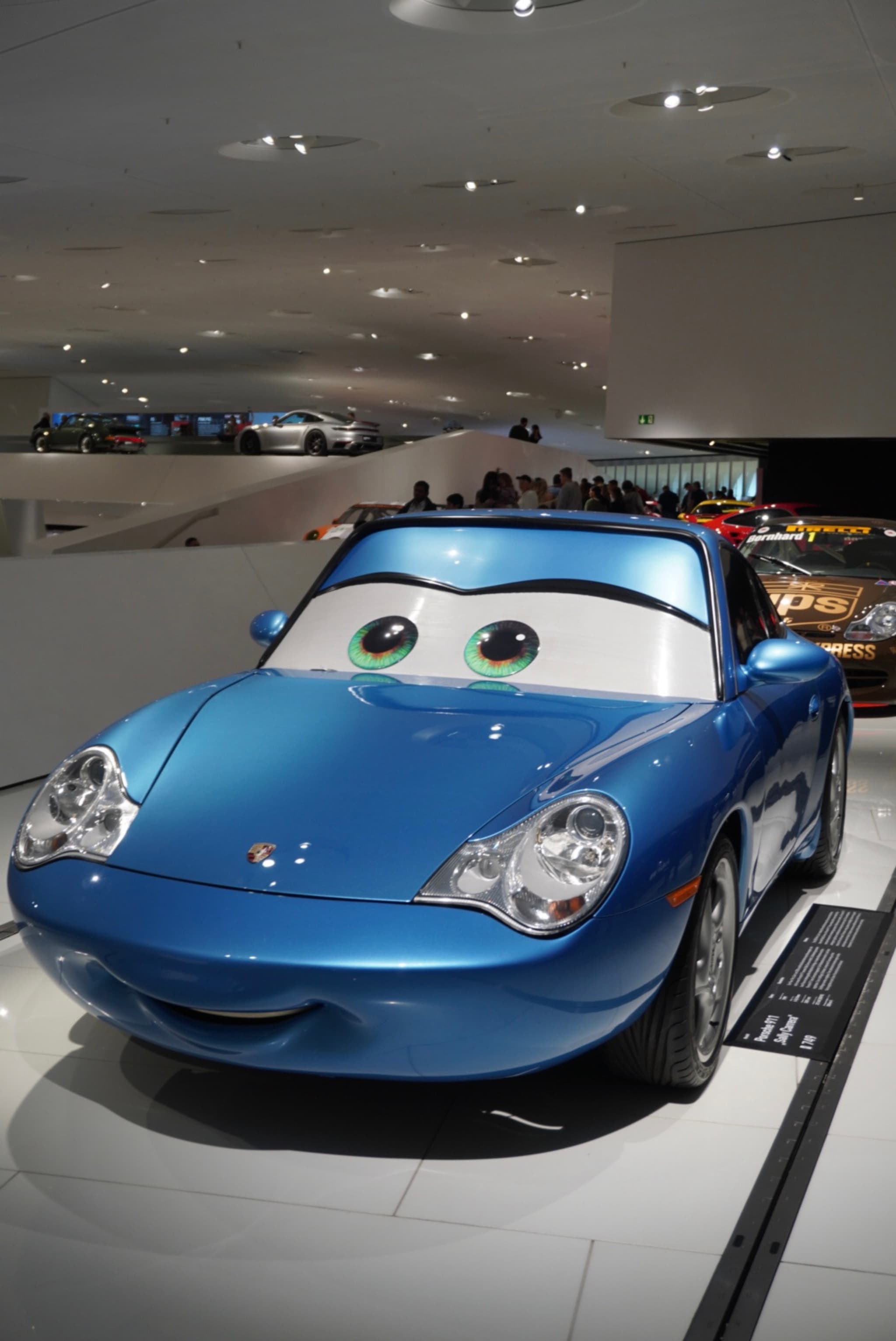 blue sally carrera character car displayed on white floor inside porsche museum under bright ceiling lights with blurred visitors in background