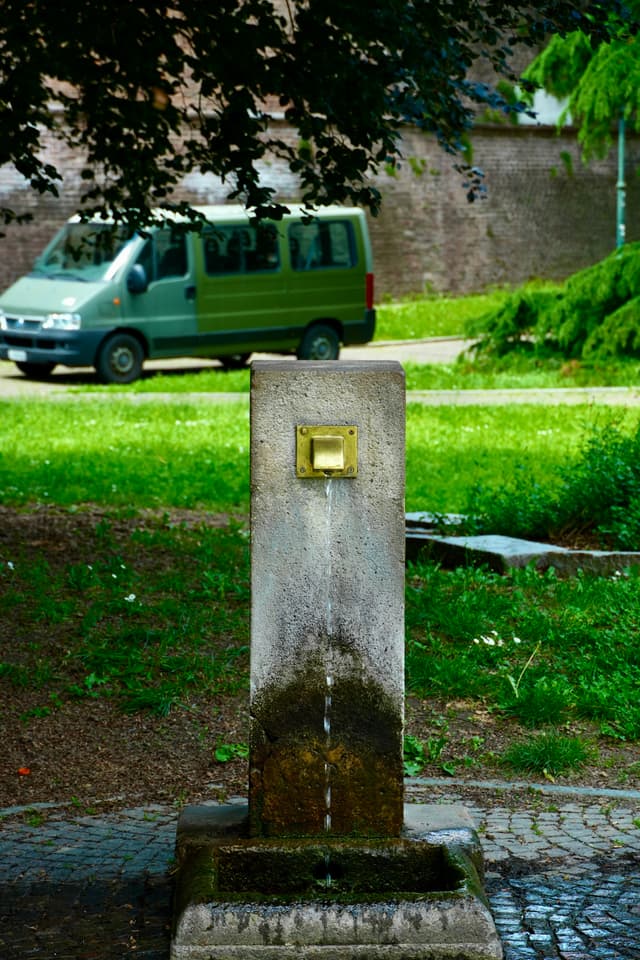 stone water fountain stands in a park with a green van parked in the background under bright daylight on a summer afternoon