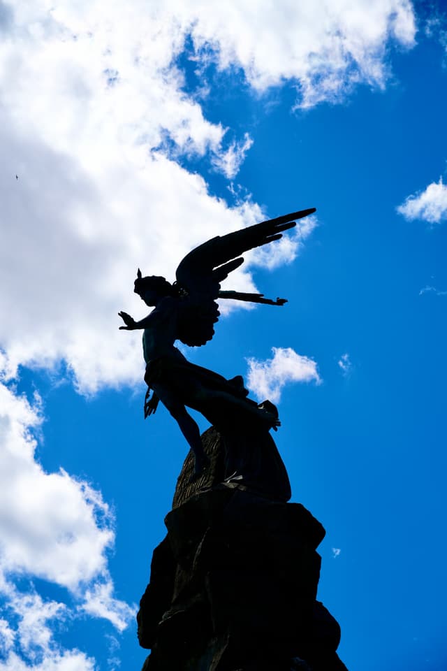 bronze angel statue silhouette against a bright blue sky with white clouds, captured from below during a sunny afternoon in a public park