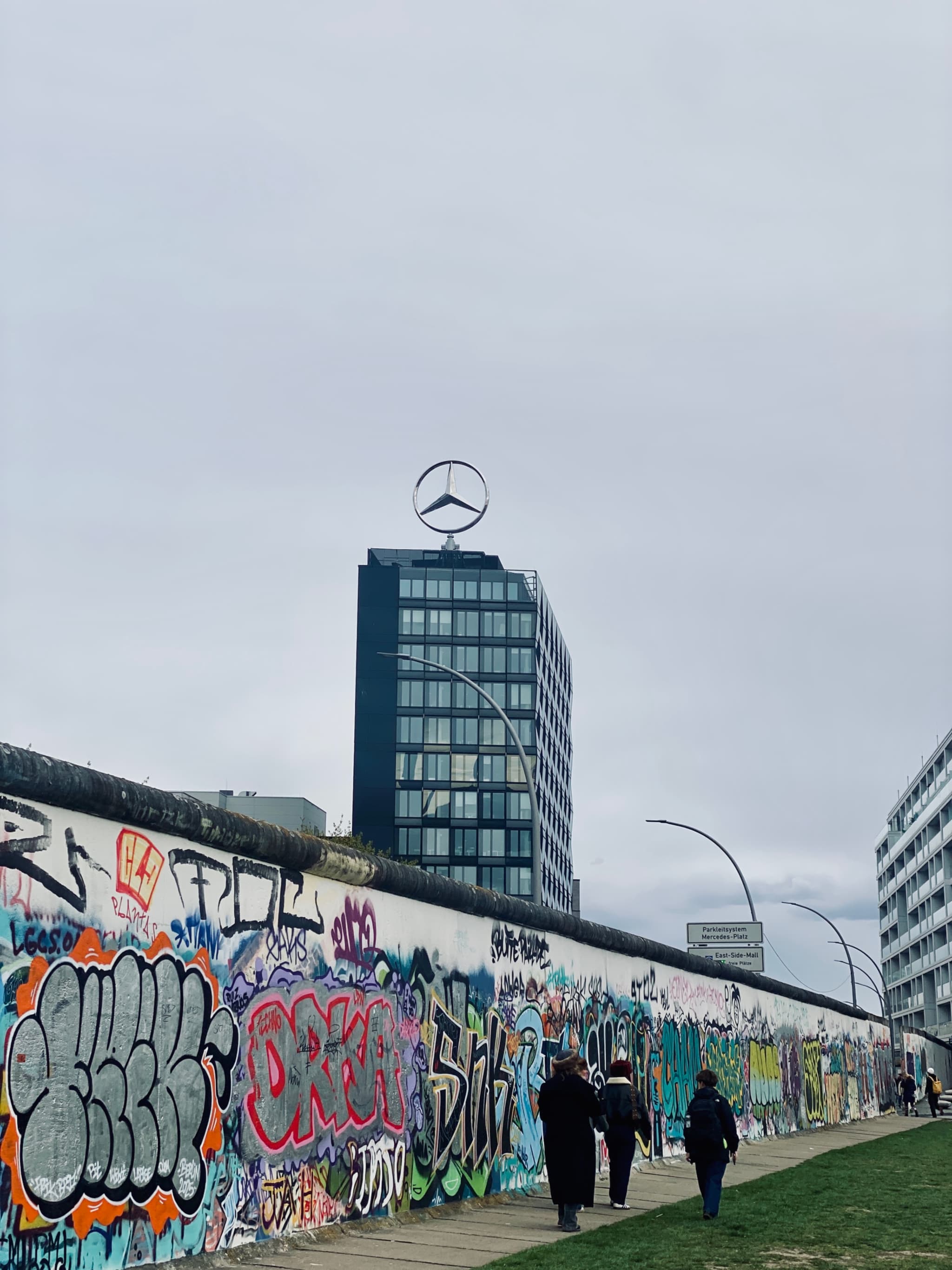 people walk past the graffiti-covered berlin wall under a gray sky with the mercedes-benz building visible in the background on a cloudy day