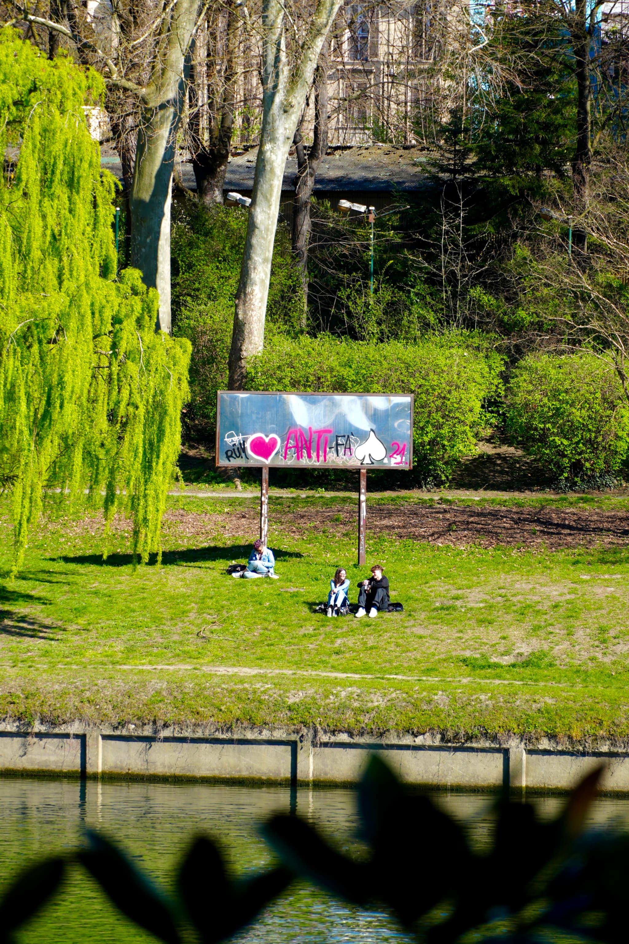 people sit on green grass near a canal under bright sunlight, framed by trees and a metal billboard with pink heart graffiti