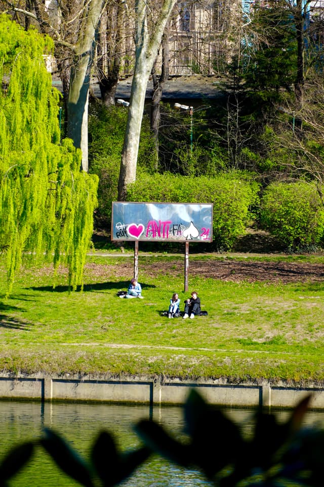 people sit on green grass near a canal under bright sunlight, framed by trees and a metal billboard with pink heart graffiti