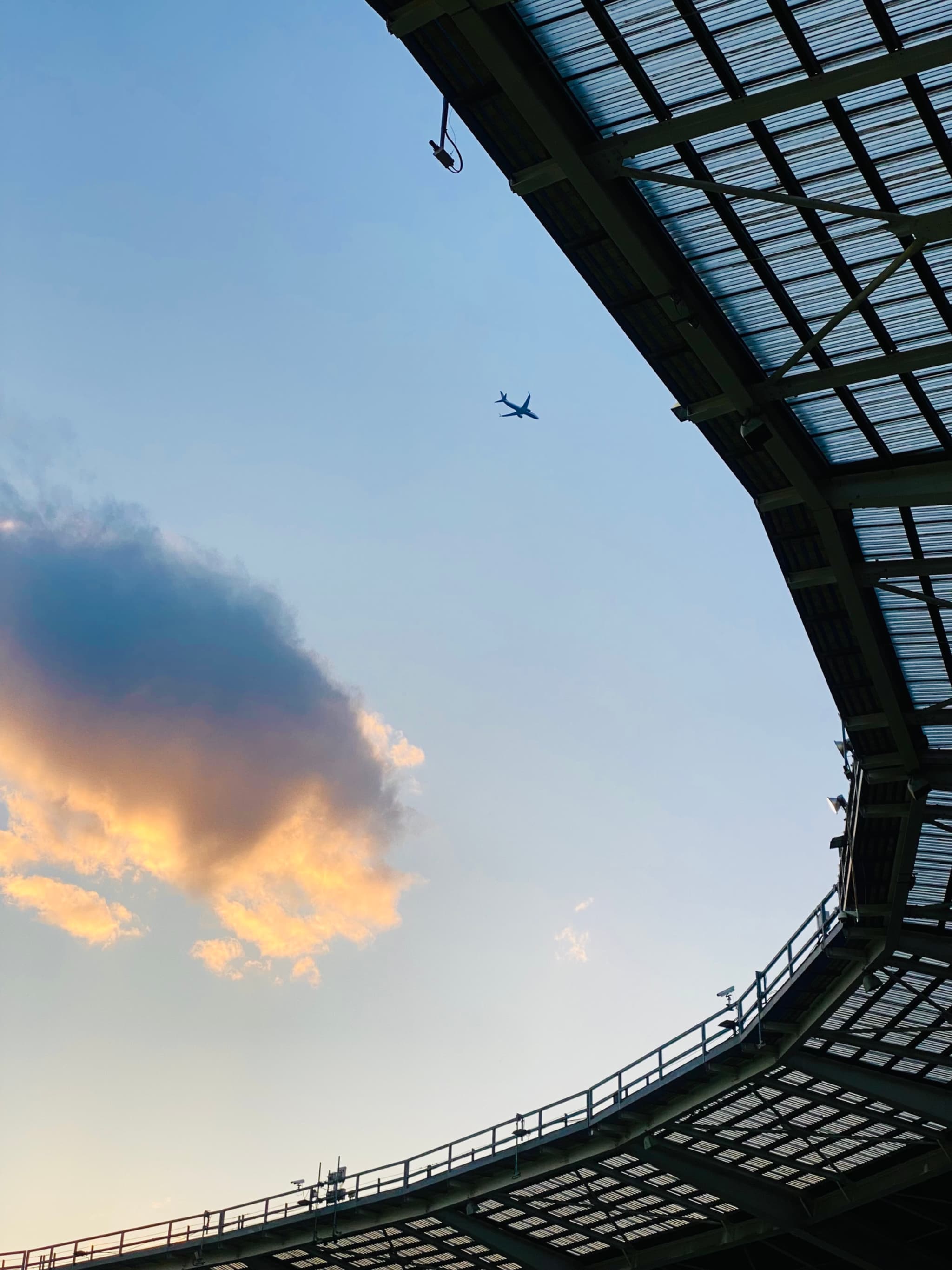 small airplane flies over a stadium roof against a clear blue sky with a single orange cloud during late afternoon light