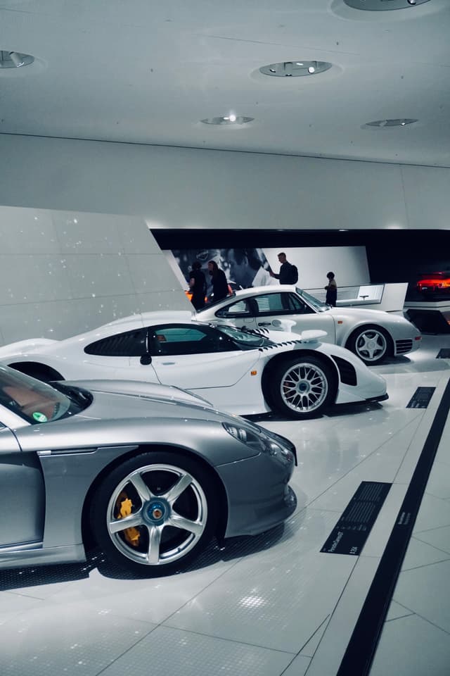 silver and white porsche sports cars parked on a white floor inside a bright automotive museum with visitors in the background