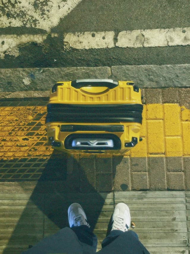 bright yellow suitcase sits on a textured city sidewalk under flat daylight, viewed from above with white sneakers in the frame