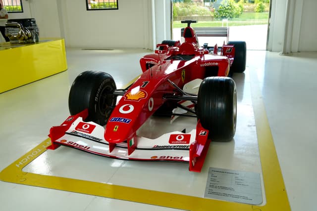 red ferrari formula one car parked on a white garage floor under bright indoor lighting with yellow painted safety lines visible