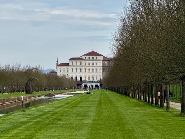 white palace architecture stands at the end of a long green lawn under a cloudy sky with rows of bare trees