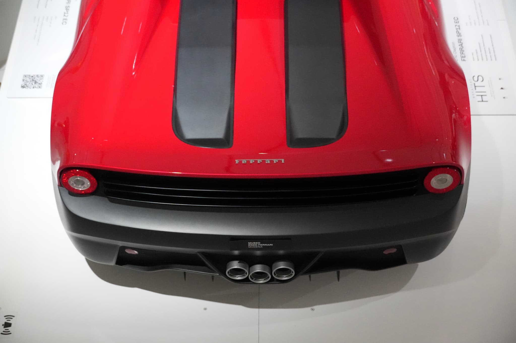 overhead view of a red ferrari sports car with black racing stripes and triple exhaust pipes parked in a bright white showroom