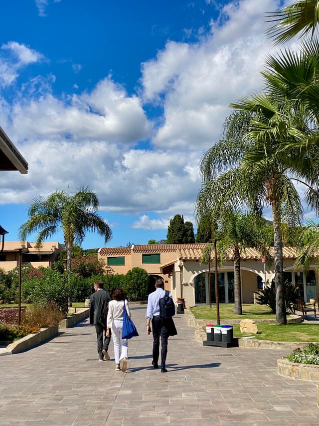 three people walk along a paved path toward a stucco building under a bright blue sky with palm trees and scattered clouds