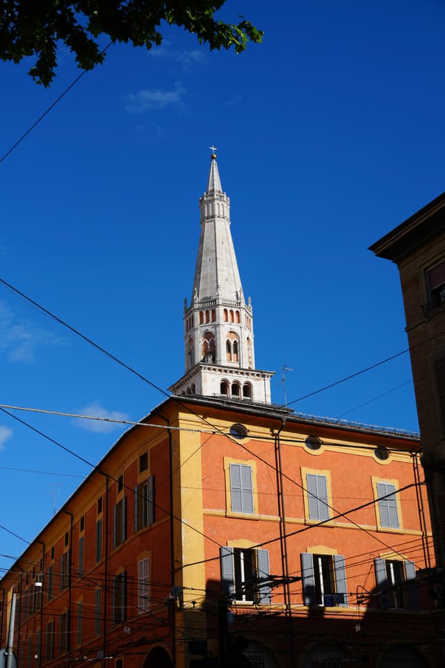 a stone church spire rises above an orange building in modena under a clear blue sky with overhead power lines crossing the frame