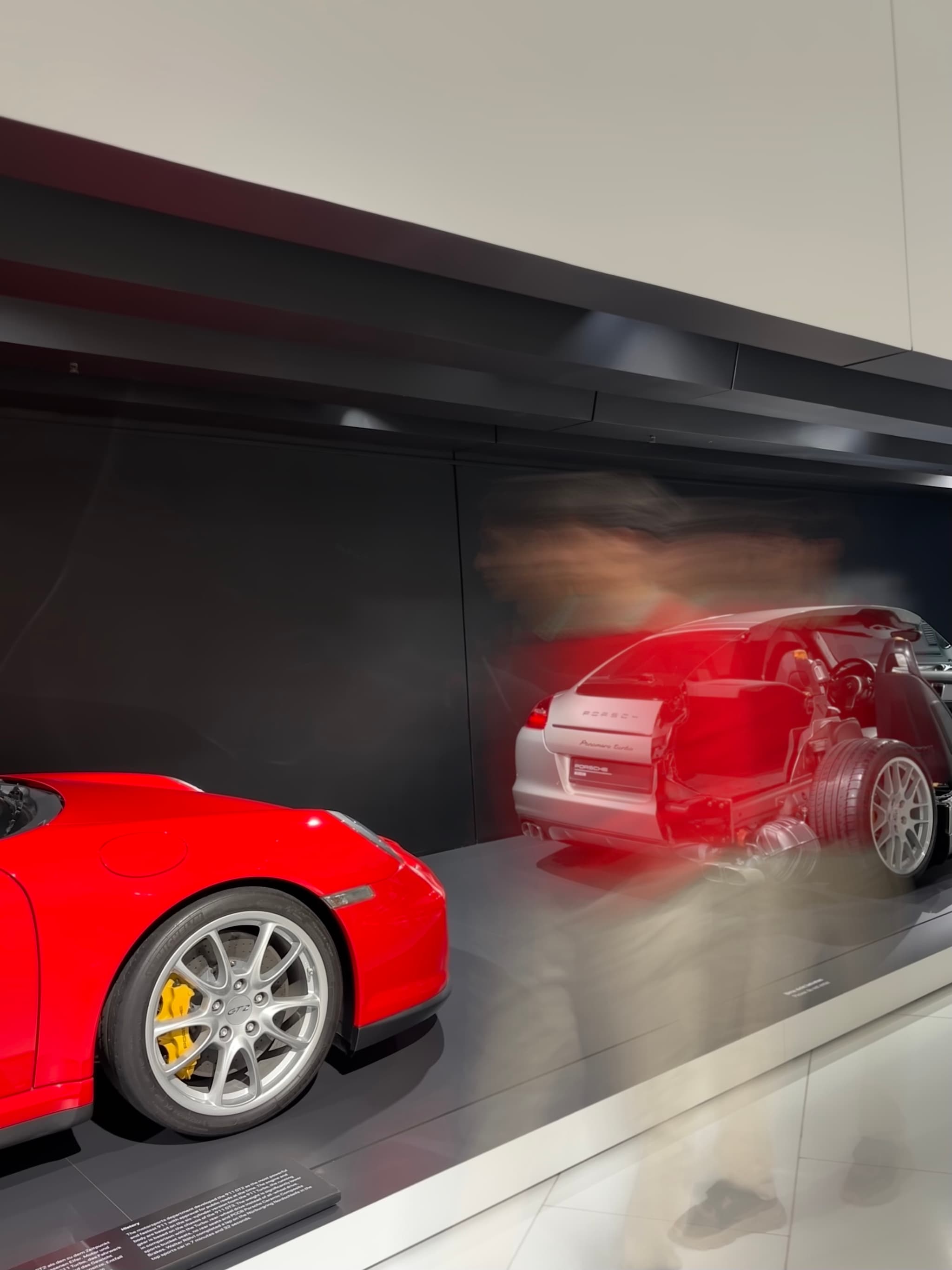 bright red sports car parked in a dark museum gallery with a motion-blurred vehicle passing by under artificial overhead lighting