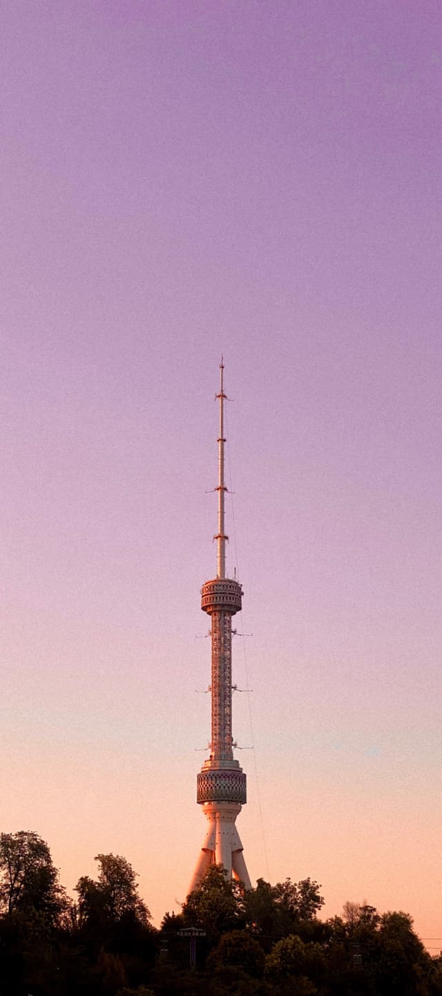 Tashkent TV Broadcasting Tower stands tall against a serene pastel sunset sky, casting a peaceful, minimalist silhouette above the lush green city trees
