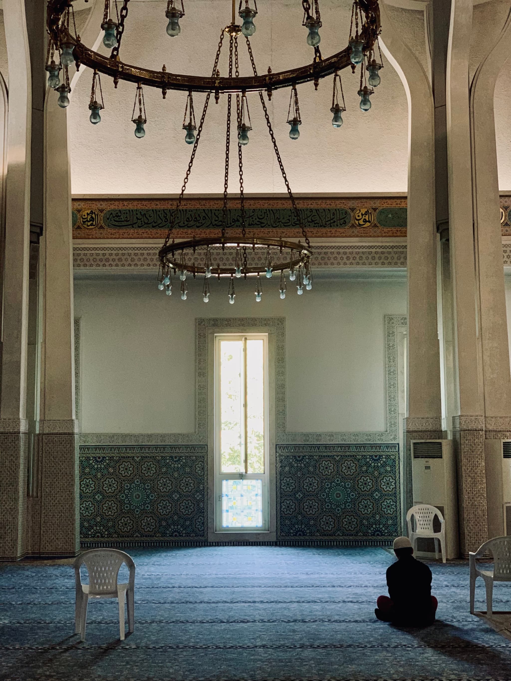 a person sits on a blue carpet inside a mosque under large metal chandeliers with soft daylight entering through a central window