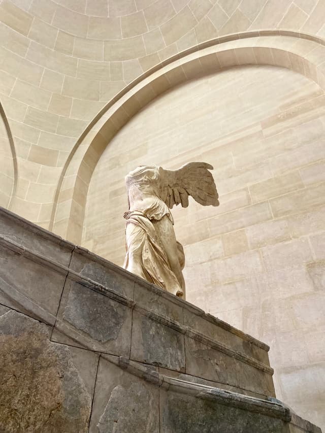 winged victory of samothrace marble statue stands on a stone staircase inside the louvre museum under soft natural overhead light