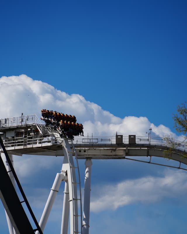 riders crest a white steel roller coaster against a bright blue sky with white clouds during a sunny afternoon at the park