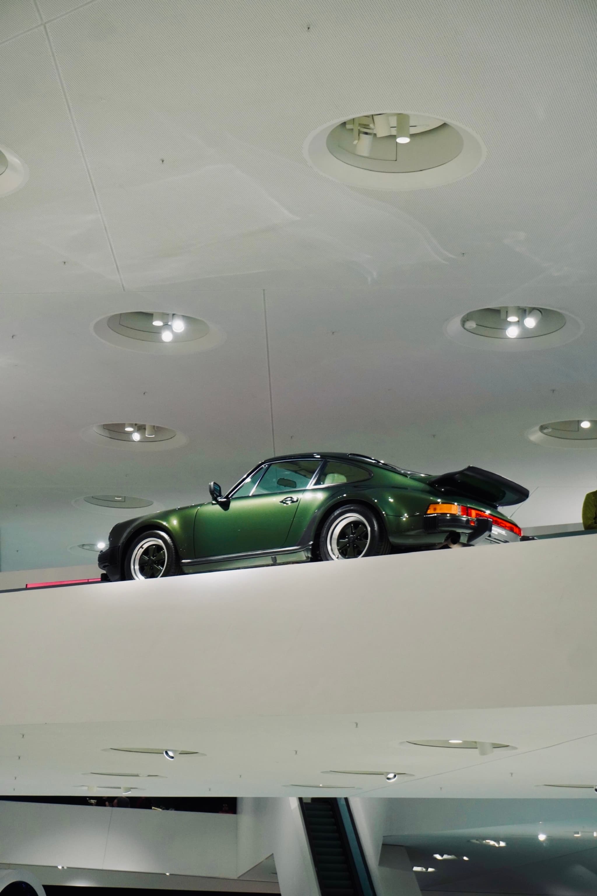 dark green porsche 911 parked on a white museum ledge under bright recessed ceiling lights in a minimalist automotive exhibition space