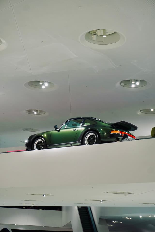 dark green porsche 911 parked on a white museum ledge under bright recessed ceiling lights in a minimalist automotive exhibition space