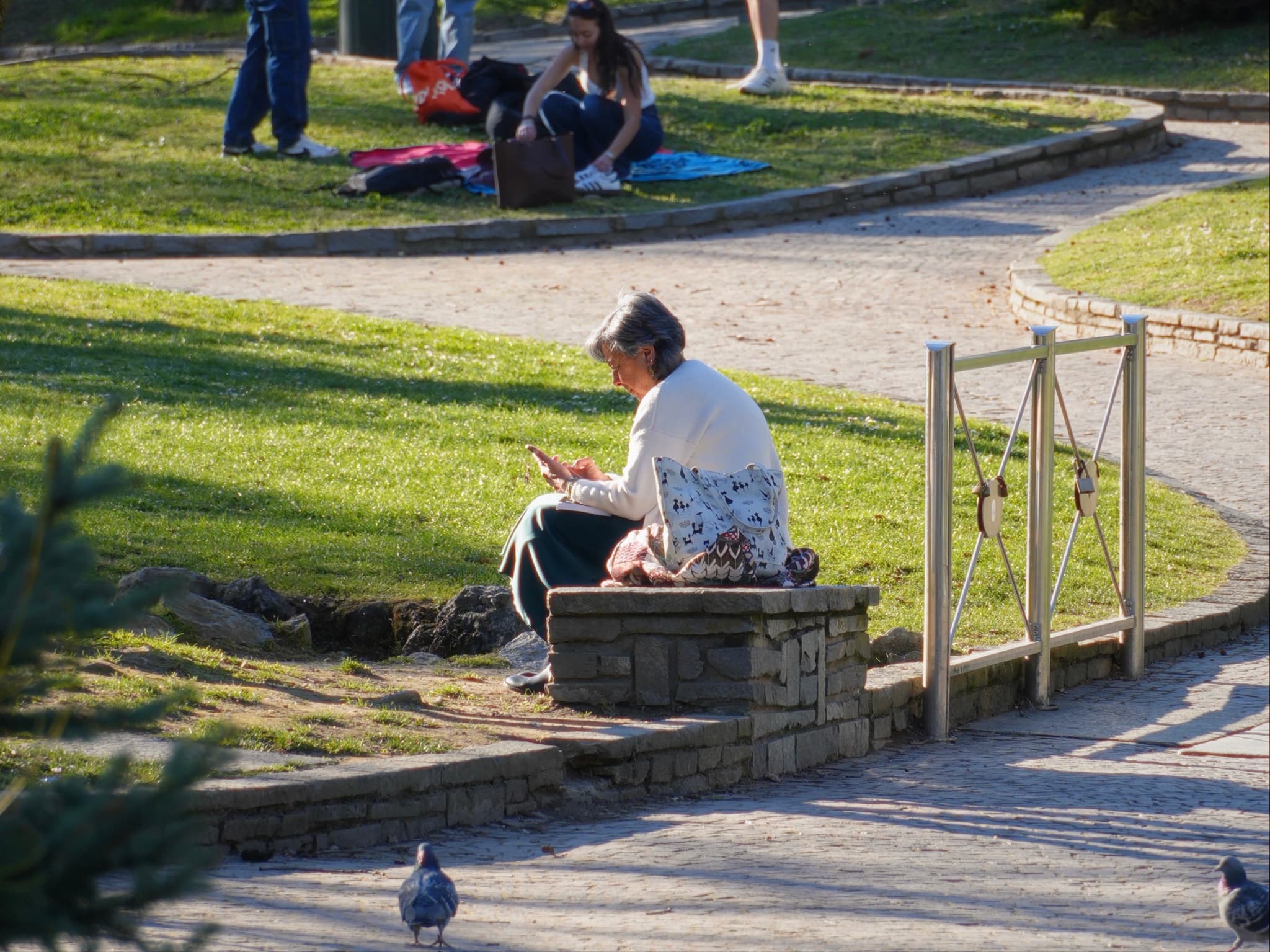 a woman sits on a stone wall in a public park during golden hour, with people relaxing on the grass nearby