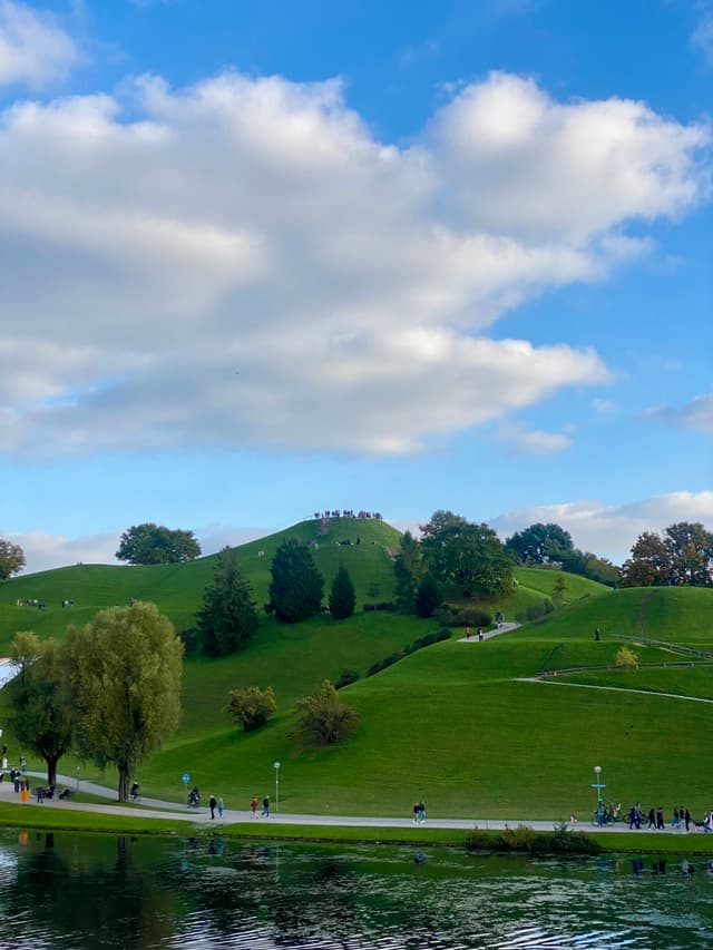 people walk along a path near green hills and a pond under a bright blue sky with white clouds in munich