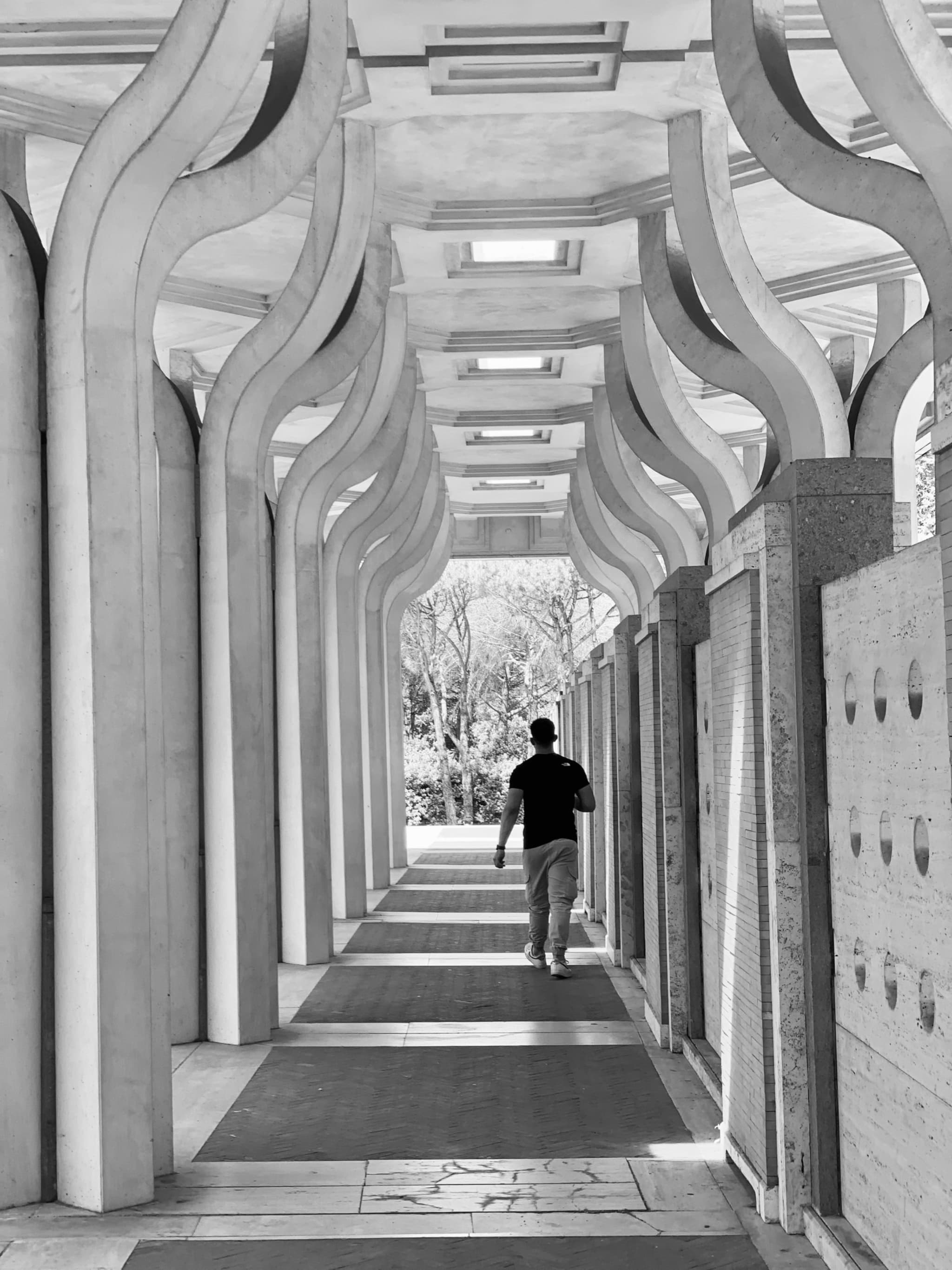person walking through a concrete colonnade with curved pillars, captured in black and white with high contrast and natural daylight
