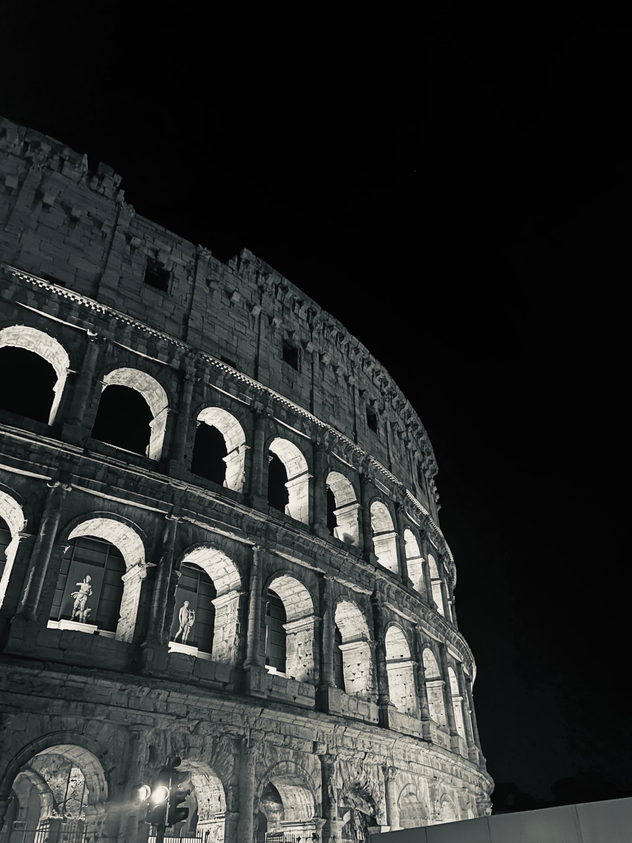 black and white low angle view of the roman colosseum at night with artificial lights highlighting the stone arches and columns